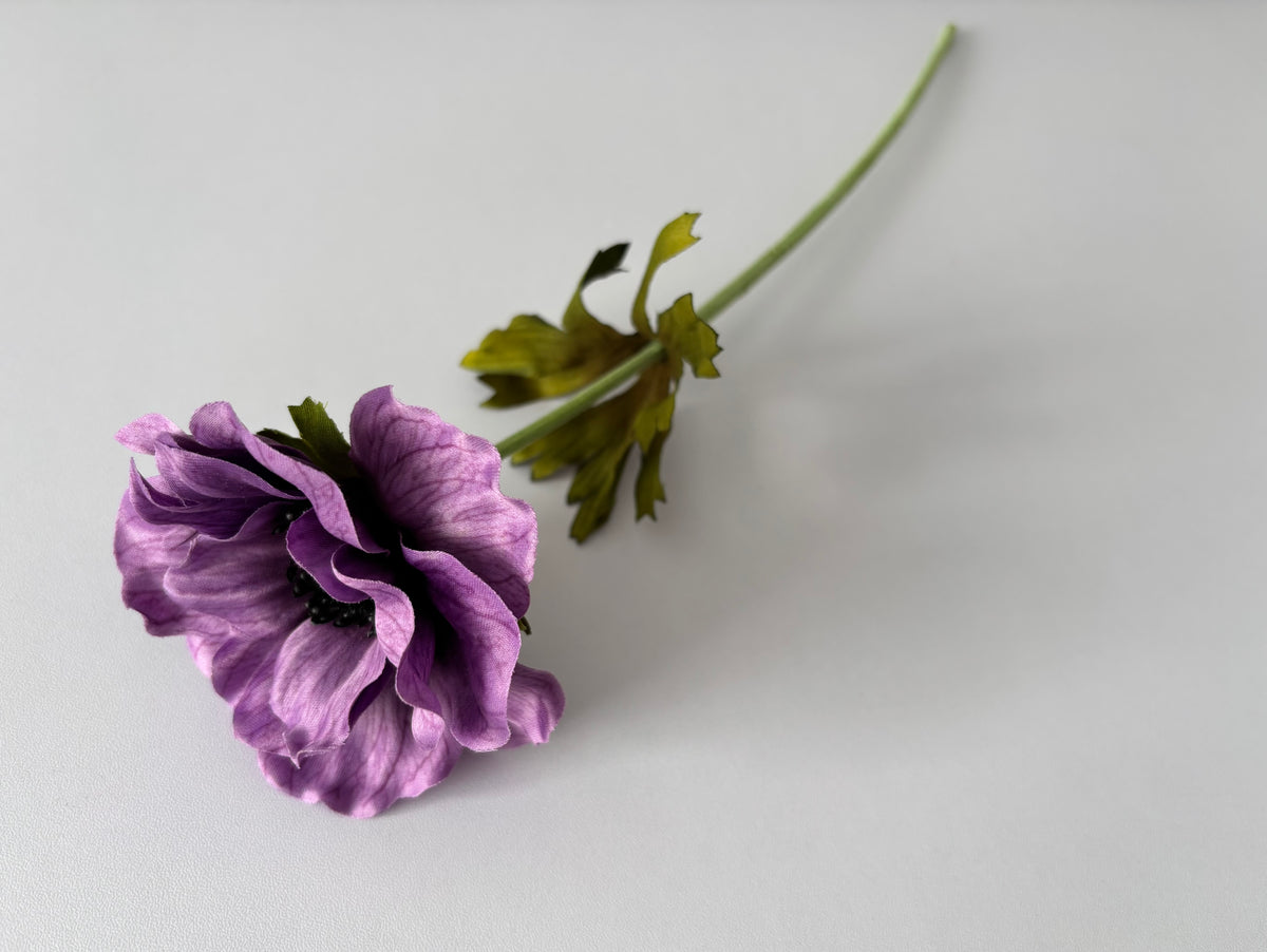 Full length flat lay shot of an artificial purple silk anemone single stem lying diagonally showing the complete 32cm fabric wrapped wired green stem with bright green deeply serrated detachable foliage leaves and an open purple anemone flower head with dark black centre drooping naturally to the left against a plain light grey background