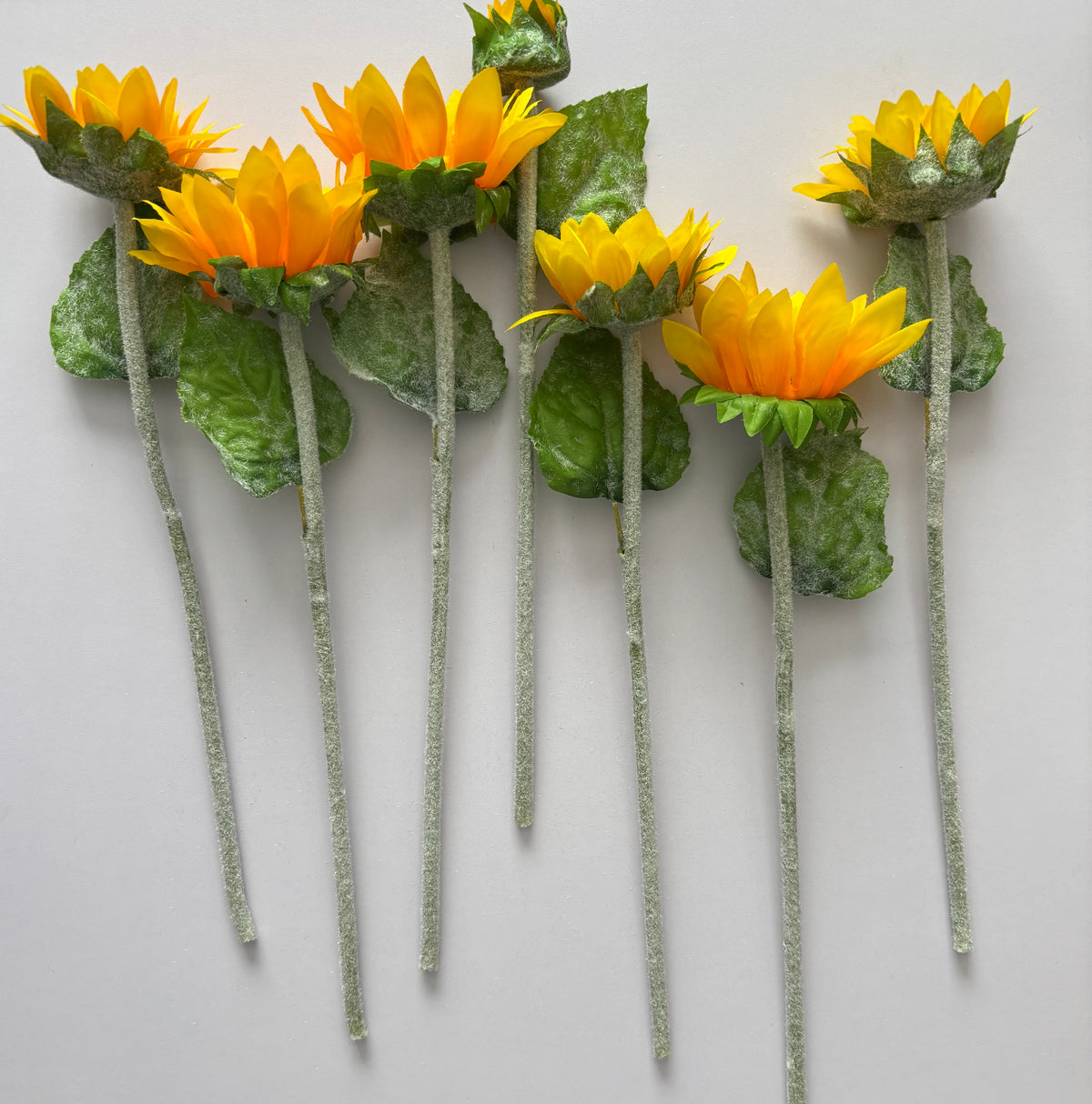 Seven artificial sunflower stems laid flat in a neat row showing the full mixed size golden yellow flower heads with dark brown centres and lush green foliage with glittery silver textured stems on a plain white background