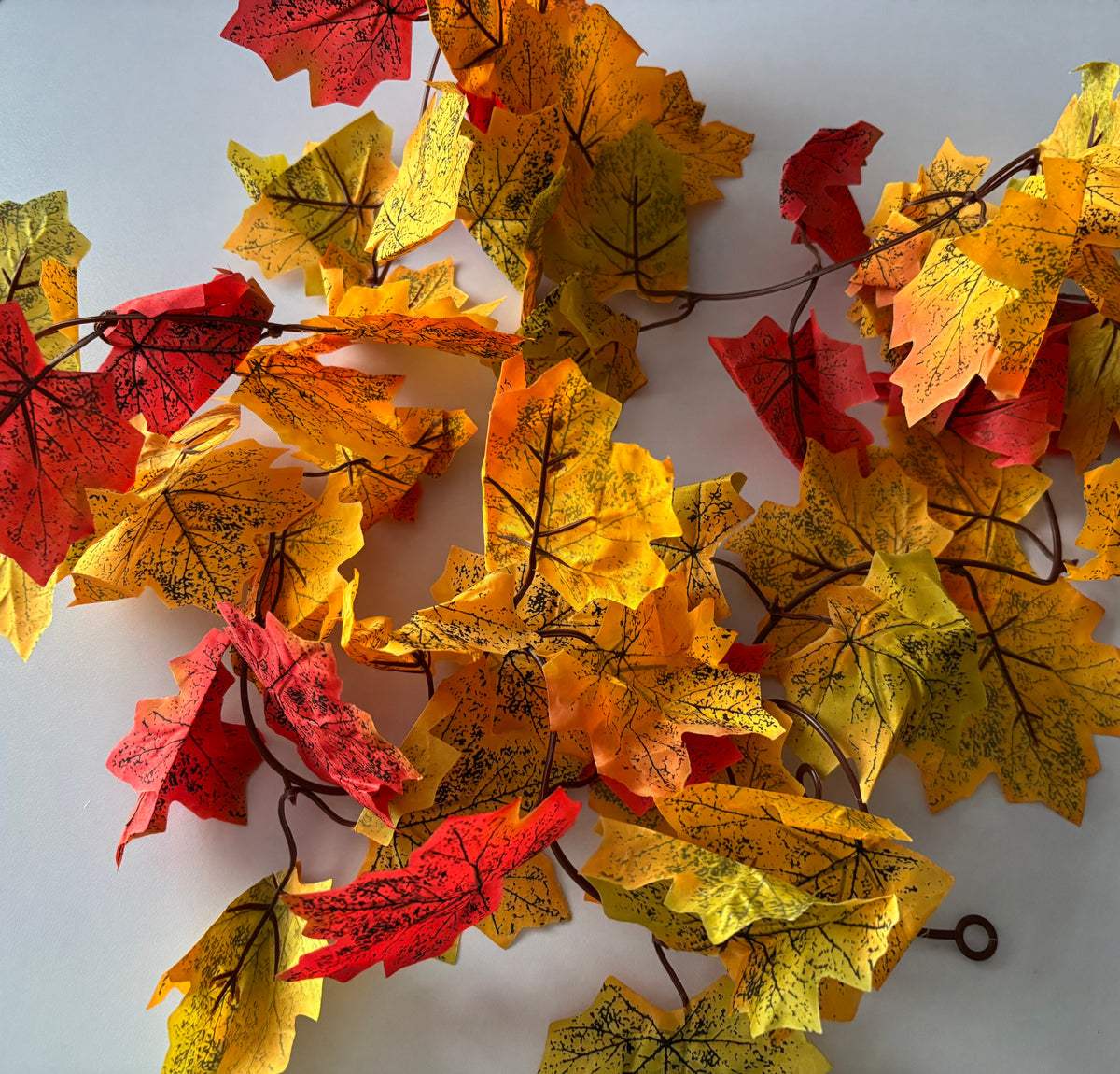 Close up flat lay shot of an autumn maple leaf garland showing multiple detachable double silk maple leaf sections in rich yellow, orange, red and green tones with detailed black leaf vein markings spread across a plain light grey background with brown plastic garland strand and hanging hoop visible at the bottom right