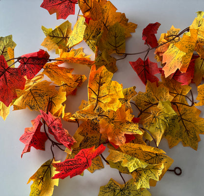 Close up flat lay shot of an autumn maple leaf garland showing multiple detachable double silk maple leaf sections in rich yellow, orange, red and green tones with detailed black leaf vein markings spread across a plain light grey background with brown plastic garland strand and hanging hoop visible at the bottom right