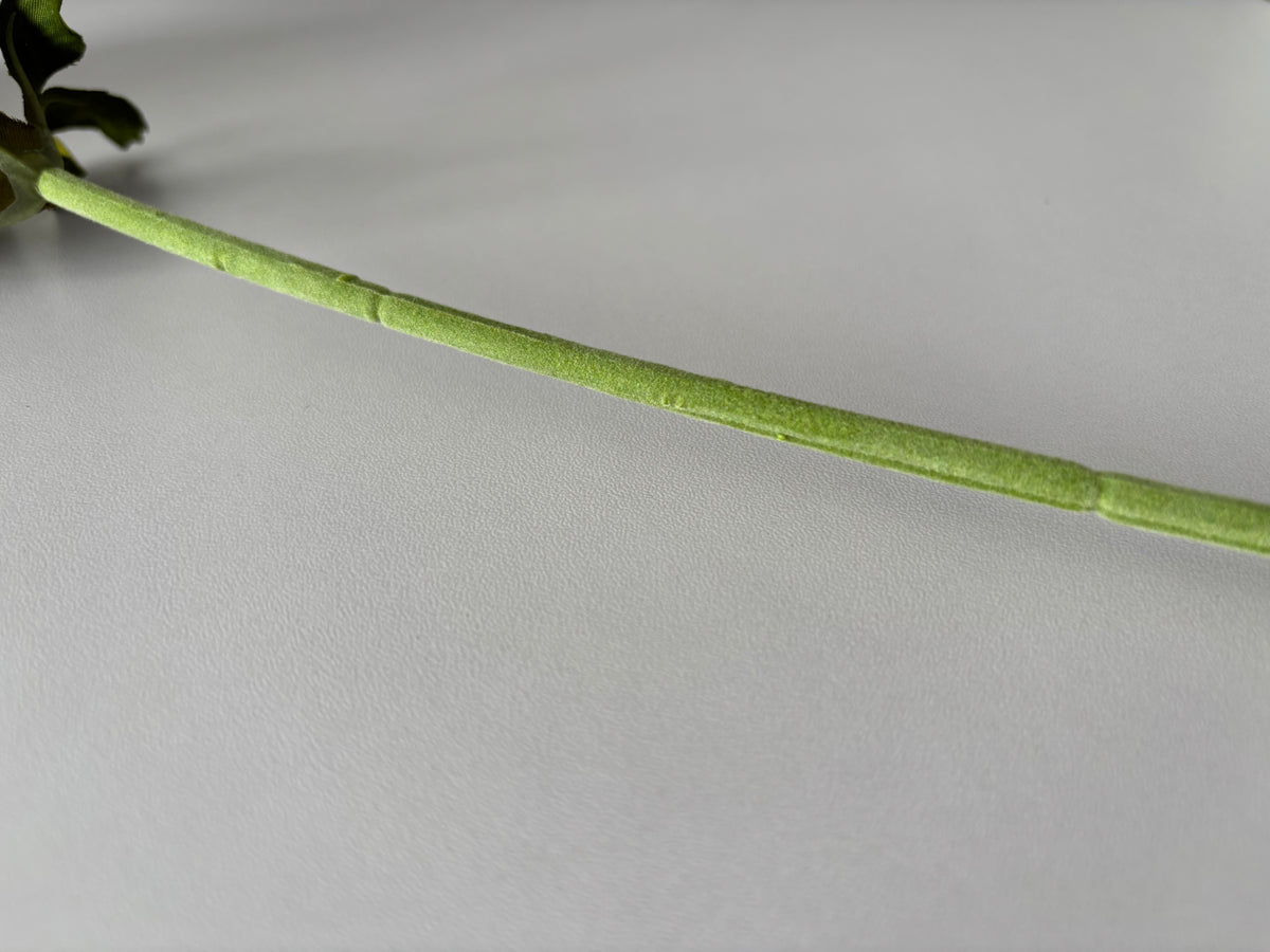 Close up diagonal shot of an artificial purple silk anemone stem showing the soft fabric wrapped wired green stem with bright green detachable serrated foliage leaves attached at the top visible against a plain light grey background