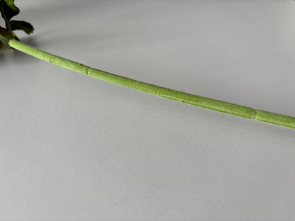 Close up diagonal shot of an artificial purple silk anemone stem showing the soft fabric wrapped wired green stem with bright green detachable serrated foliage leaves attached at the top visible against a plain light grey background