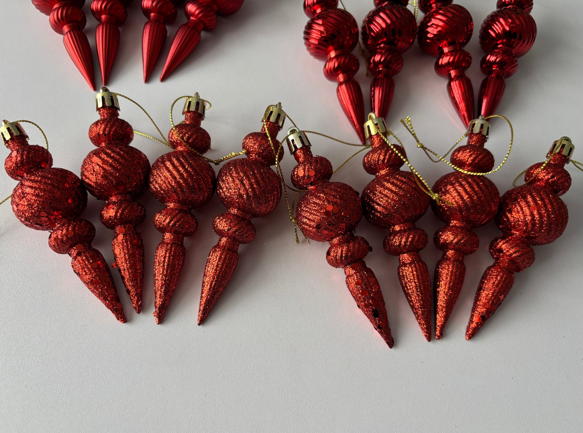 Overhead flat lay of red Christmas spindle baubles fanned out in two rows showing metallic and glittered finishes with ribbed spherical tops, beaded middles and elongated pointed drop bases on a plain light grey background