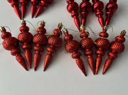 Overhead flat lay of red Christmas spindle baubles fanned out in two rows showing metallic and glittered finishes with ribbed spherical tops, beaded middles and elongated pointed drop bases on a plain light grey background