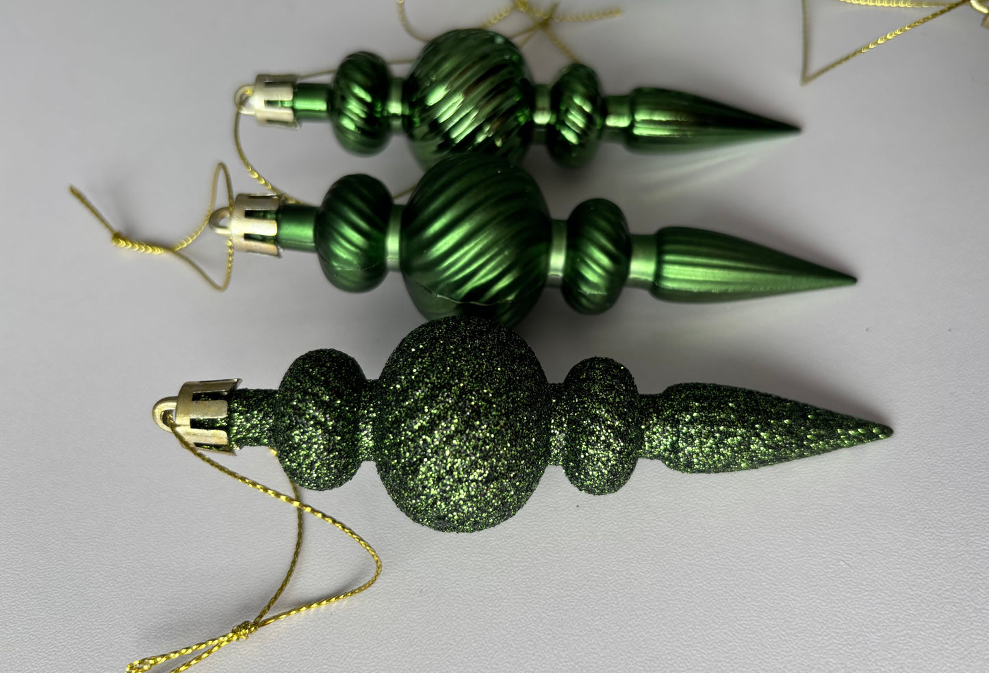 Close up of three green Christmas spindle baubles showing metallic and glittered finishes side by side with ribbed spherical tops, beaded middles and elongated pointed drop bases with gold hanging hooks on a plain white background
