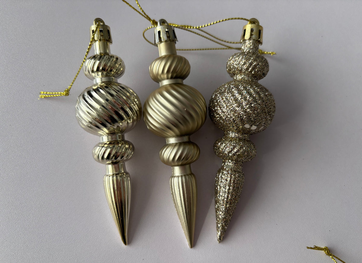 Three champagne gold Christmas spindle baubles side by side showing matte, metallic and glittered finishes with ribbed bulbous spherical tops, twisted ribbed middles and elongated pointed drop bases with gold hanging strings on a plain light grey background