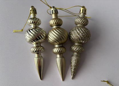 Three champagne gold Christmas spindle baubles side by side showing matte, metallic and glittered finishes with ribbed bulbous spherical tops, twisted ribbed middles and elongated pointed drop bases with gold hanging strings on a plain light grey background