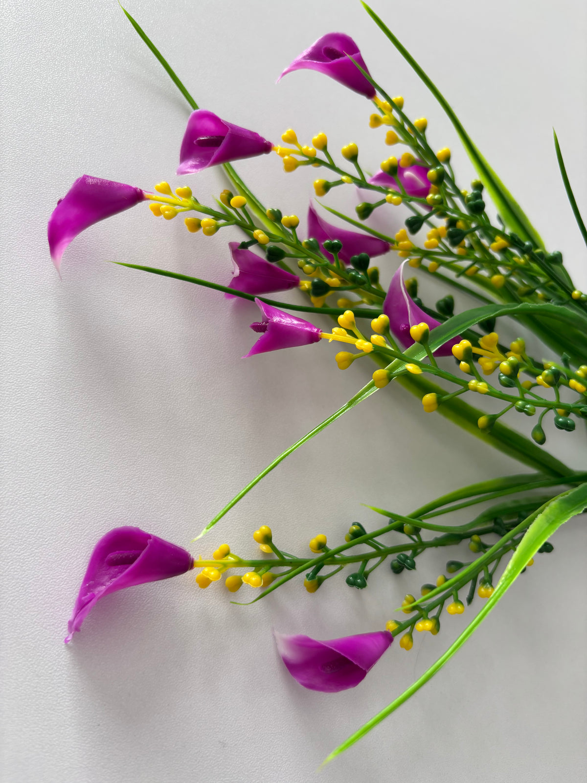 Upright portrait shot of an artificial pink small calla lily foliage bunch from a low close angle showing multiple small bright magenta pink calla lily flower heads drooping naturally with dense clusters of bright yellow berry pick stems and tall thin green grass stems spreading upward against a plain light grey background