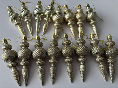Flat lay of all 16 champagne gold Christmas spindle baubles in glittered finish showing chunky glitter texture arranged in two rows with ribbed spherical tops, twisted middles and elongated pointed drop bases with gold hanging strings on a plain white background