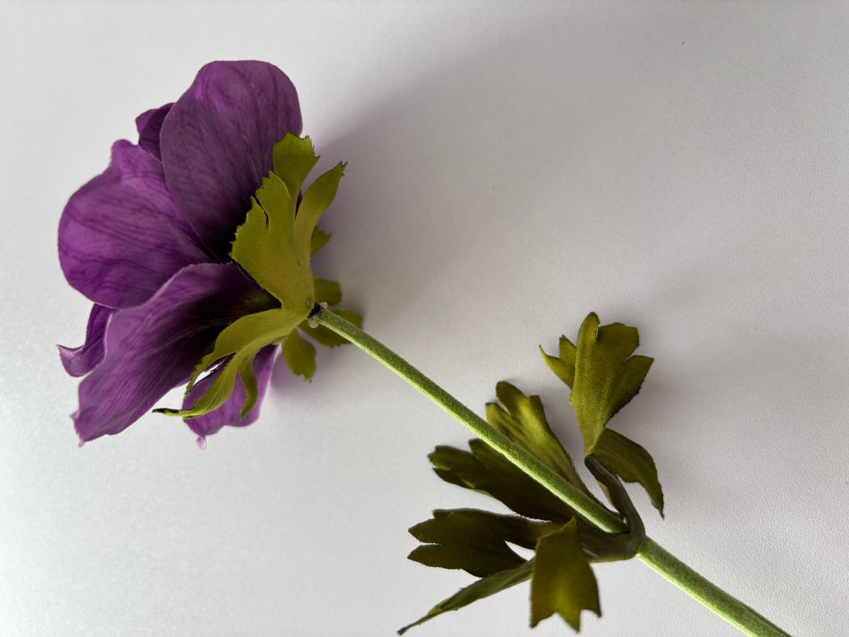 Angled side view shot of an artificial purple silk anemone single stem showing the open flower head with layered purple petals and dark black centre with two large bright green deeply serrated detachable foliage leaves spread out along the fabric wrapped green stem against a plain light background