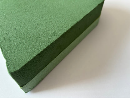 Side profile close up shot of a green heart shaped oasis wet floral foam frame showing the full 6cm depth and two layer construction with the soft porous wet foam top layer for stem insertion and the solid firm green base layer for easy handling clearly visible against a plain white background