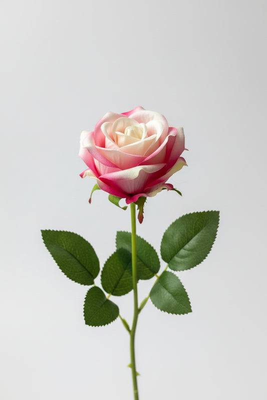 AI lifestyle image of a single artificial pink and white velvet touch rose stem showing full stem length with detachable green leaves and a soft blush pink and cream two-tone rose head on a plain neutral background