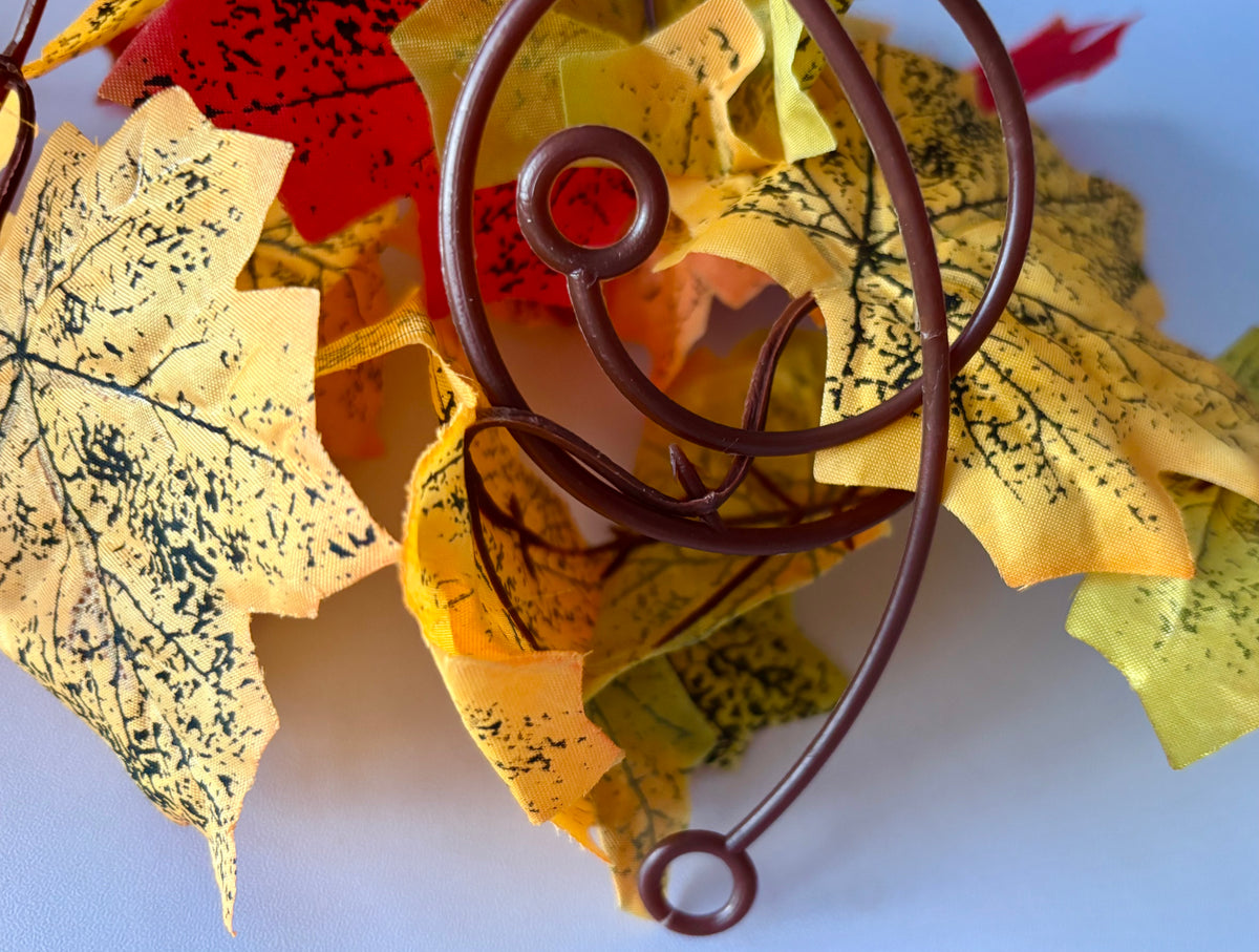 Close up detail shot of an autumn maple leaf garland 180cm showing the two brown flexible plastic hanging hoops on the brown plastic garland strand surrounded by multiple silk maple leaves in yellow, orange, red and green tones with detailed black leaf vein markings against a plain light blue background