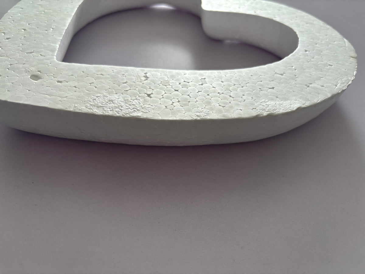 Extreme close up macro side profile shot of a white polystyrene heart wreath frame showing the full 3.5cm depth and highly detailed textured granular porous polystyrene cut edge surface with the hollow centre visible against a plain neutral grey background