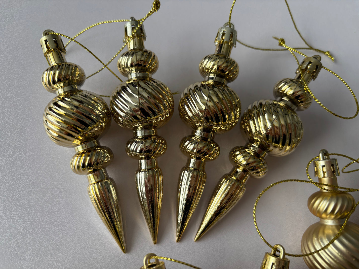 Close up of four bright gold metallic Christmas spindle baubles with highly polished ribbed spherical tops, ribbed middles and elongated pointed drop bases hanging from gold twisted cord strings on a plain light grey background