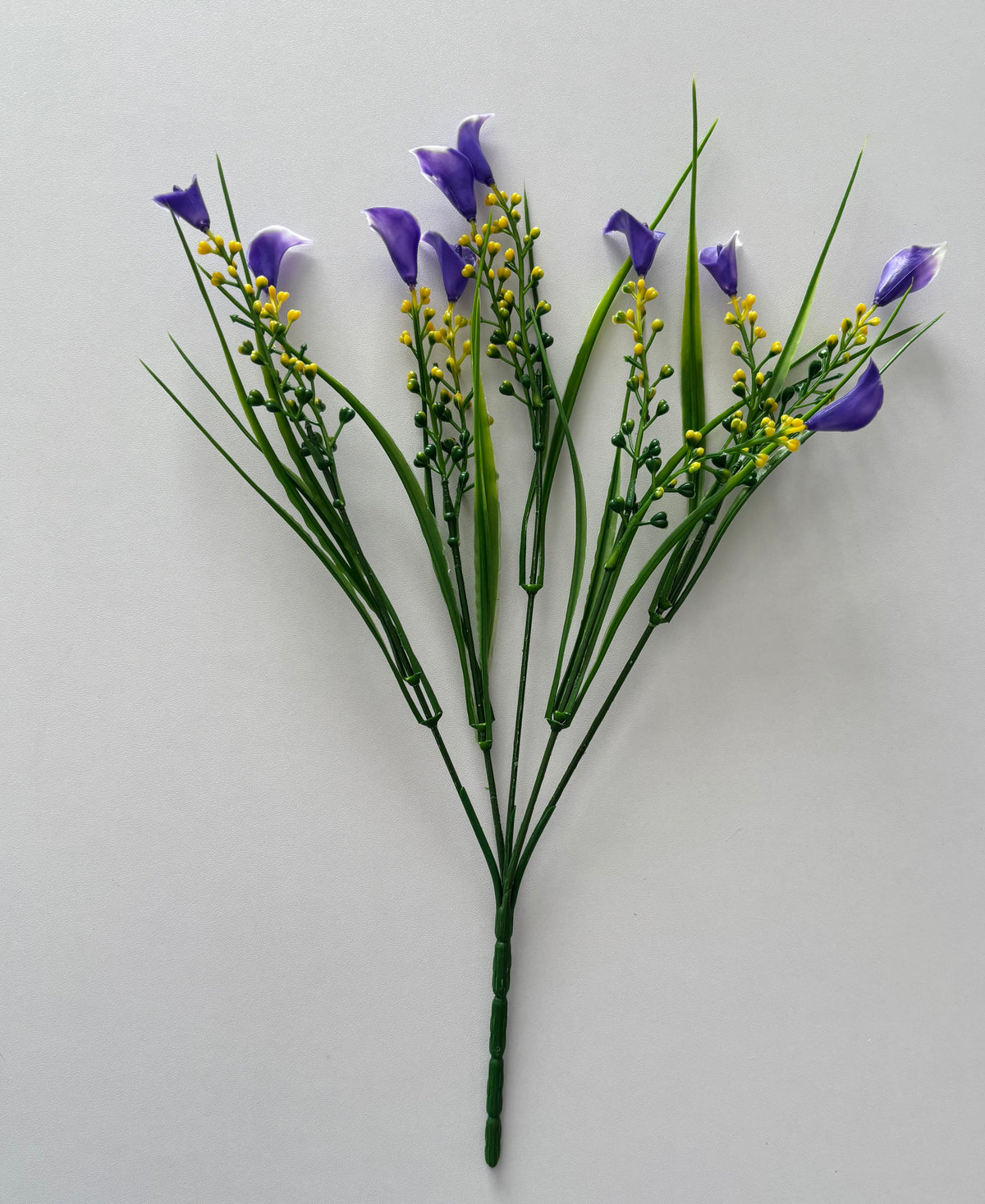 Upright portrait shot of an artificial purple small calla lily foliage bunch showing the full bunch height with five individual wired stems fanning out from a single bottom stem with small purple calla lily flower heads, tall thin green grass stems and small bright yellow berry pick stems against a plain light grey background