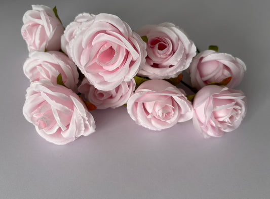 Close up overhead shot of a cluster of seven artificial blush to mid-pink silk rose heads with beautifully layered soft petals and small green foliage leaves arranged on a plain light grey background