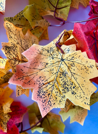 Extreme close up macro shot of a single silk maple leaf from an autumn maple leaf garland showing rich yellow, orange and pink colour tones with detailed black leaf vein markings and realistic leaf texture against a bright blue and lavender blurred background
