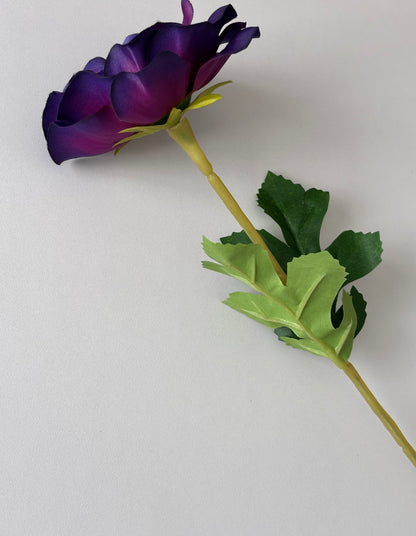 Diagonal portrait shot of an artificial purple anemone single stem showing the partially open deep purple and violet flower bud at the top with bright yellow-green calyx wired stem and lush dark green serrated foliage leaves fanned out at the bottom on a plain light background