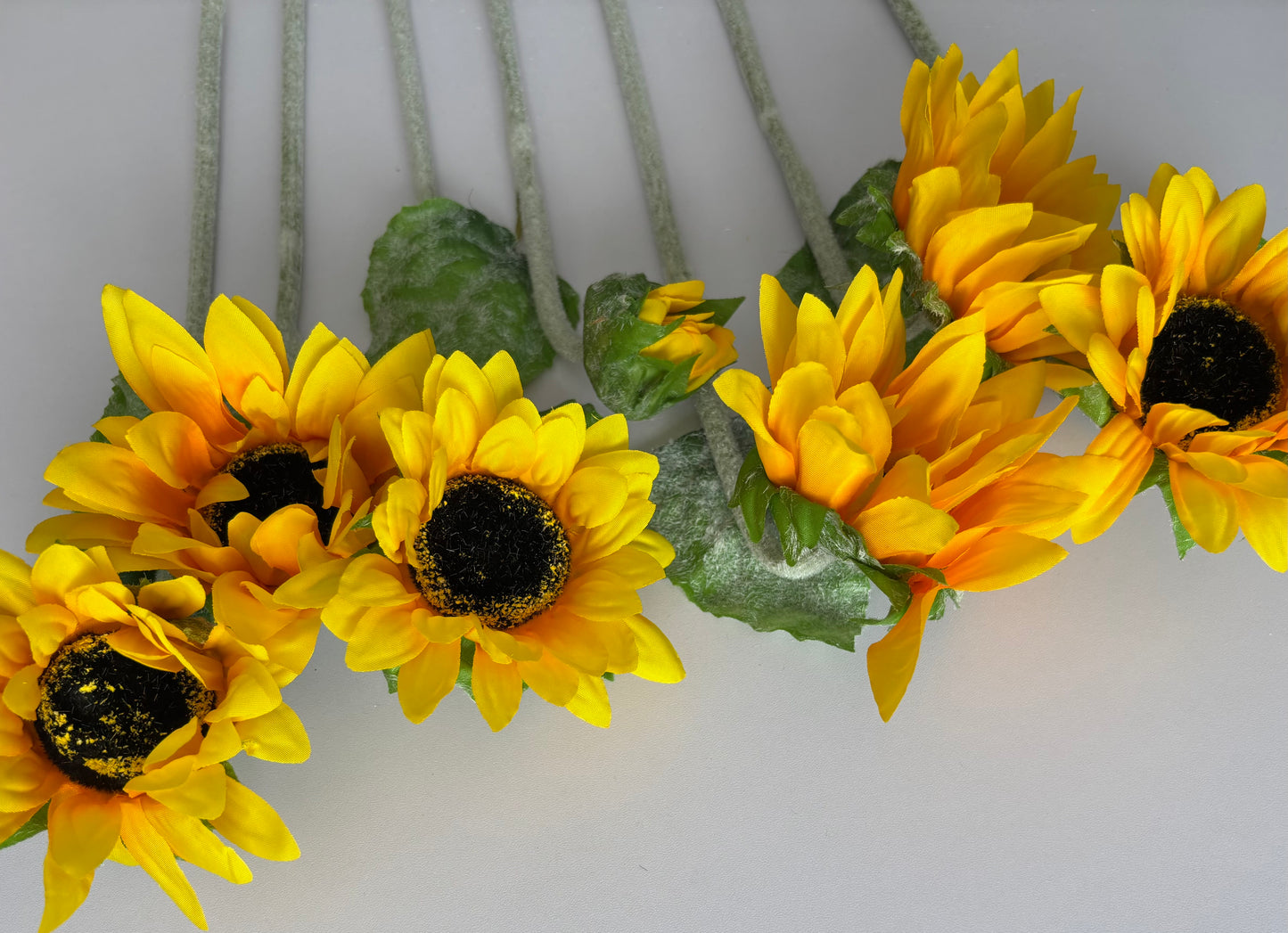 Elevated close up shot of five artificial sunflower heads showing the mixed size golden yellow petals with orange tones dark brown centres and lush green foliage fanned out with glittery silver textured stems visible on a plain white background