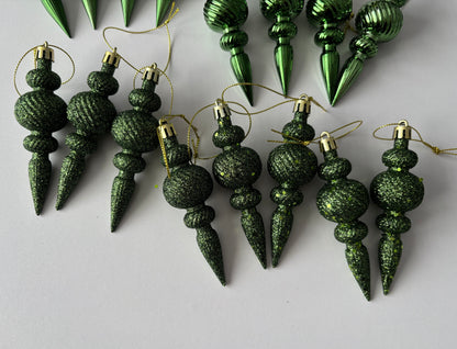Flat lay of eight green glittered Christmas spindle baubles with chunky glitter textured spherical tops, beaded middles and elongated pointed drop bases with gold hanging strings arranged on a plain white background with metallic baubles visible at the top