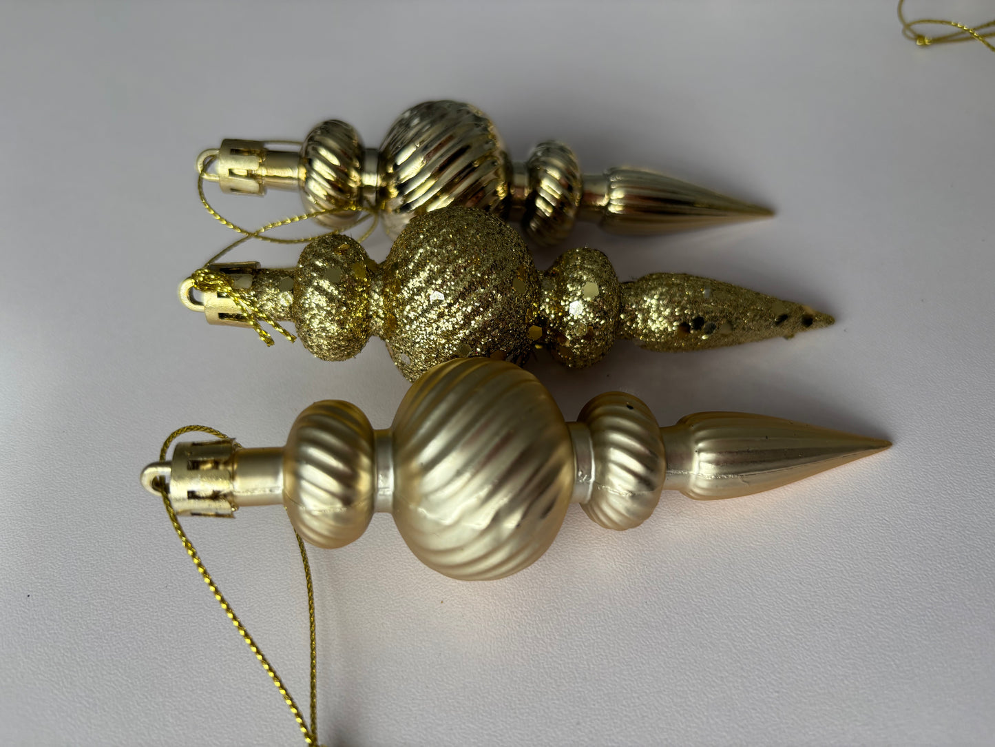 Three gold Christmas spindle baubles arranged at an angle showing glittered and matte finishes with ribbed spherical tops, beaded middles and elongated pointed drop bases with gold hanging hooks on a plain light grey background