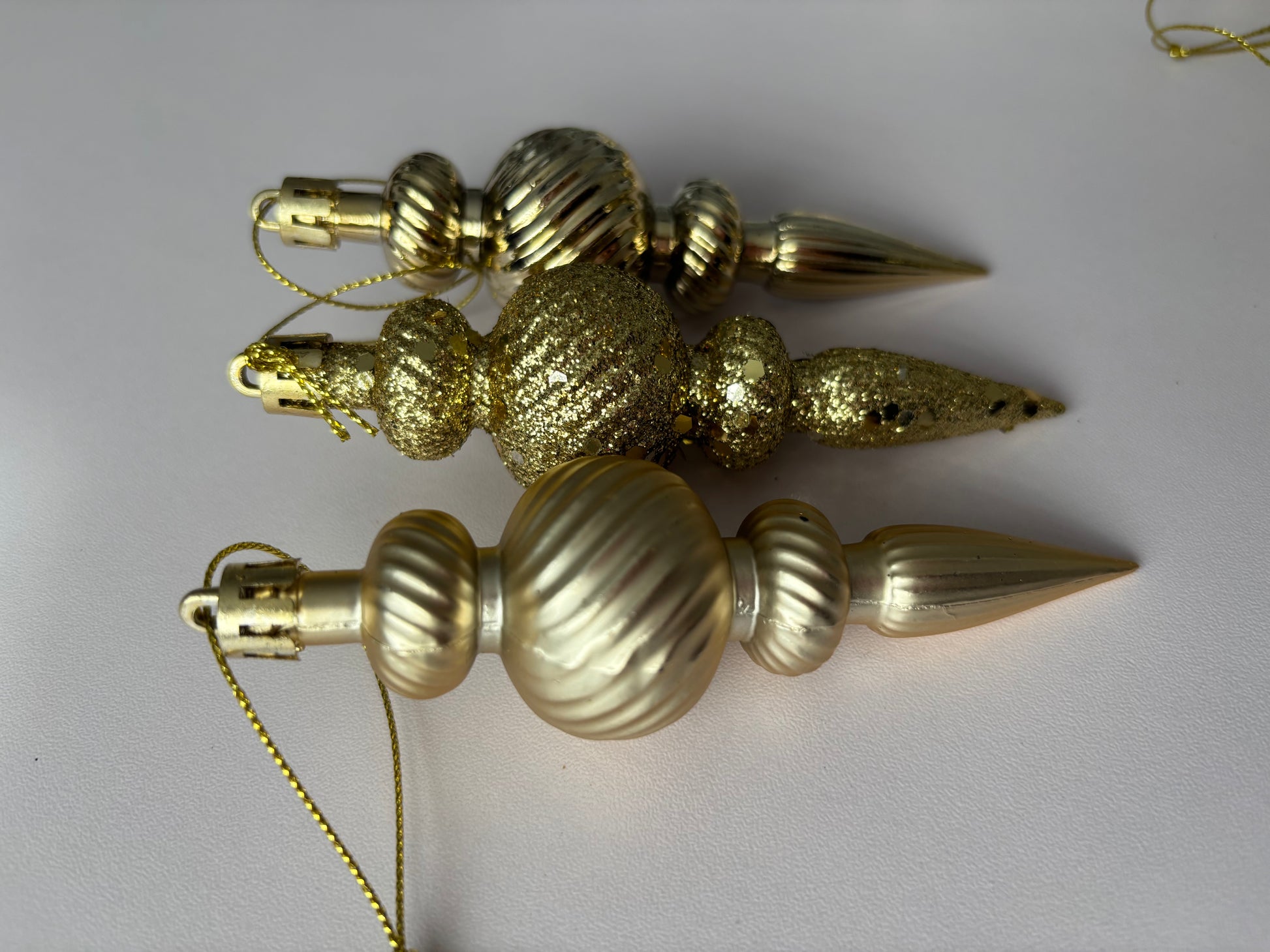 Three gold Christmas spindle baubles arranged at an angle showing glittered and matte finishes with ribbed spherical tops, beaded middles and elongated pointed drop bases with gold hanging hooks on a plain light grey background