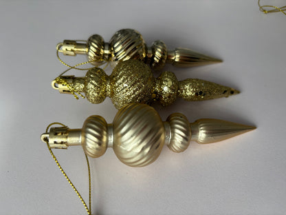 Three gold Christmas spindle baubles arranged at an angle showing glittered and matte finishes with ribbed spherical tops, beaded middles and elongated pointed drop bases with gold hanging hooks on a plain light grey background