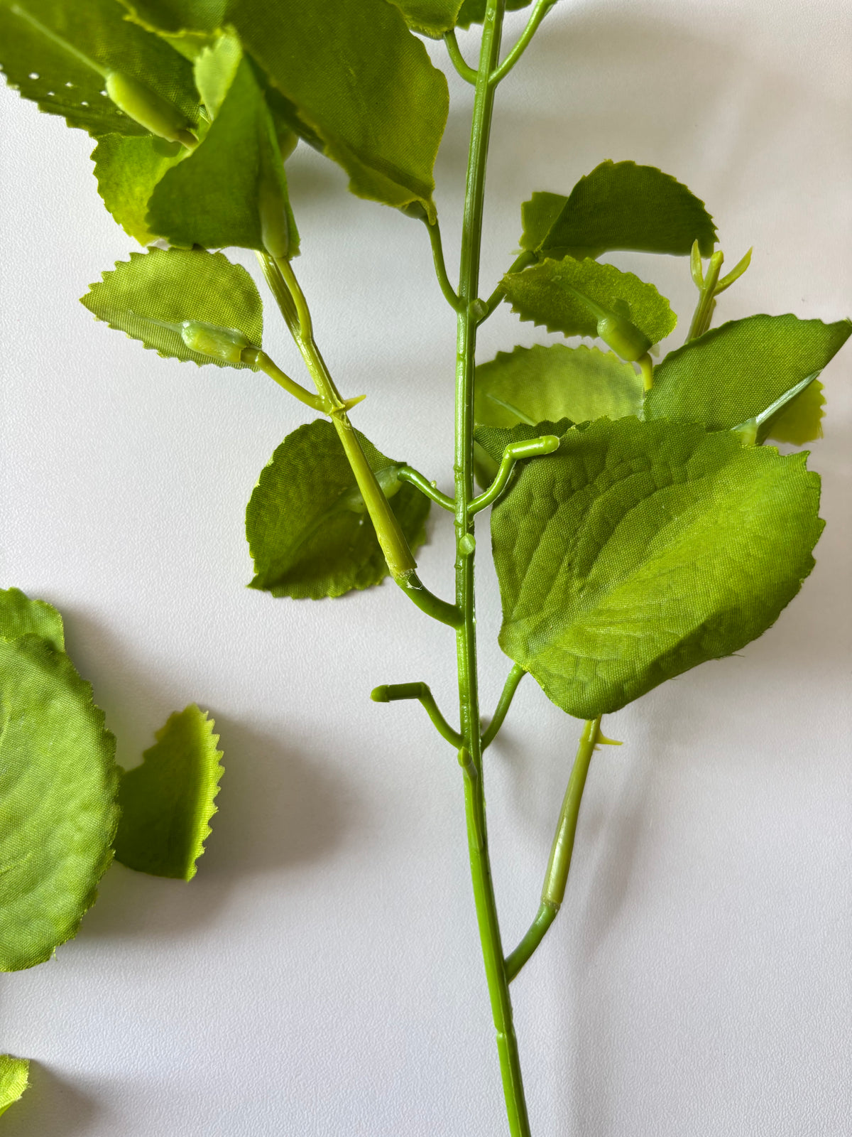 Upright close up shot of an artificial green patchouli leaf foliage spray showing the full stem height with the main wired plastic stem, two branching side stems and multiple rounded serrated green fabric silk patchouli leaves arranged naturally at varying heights against a plain white background