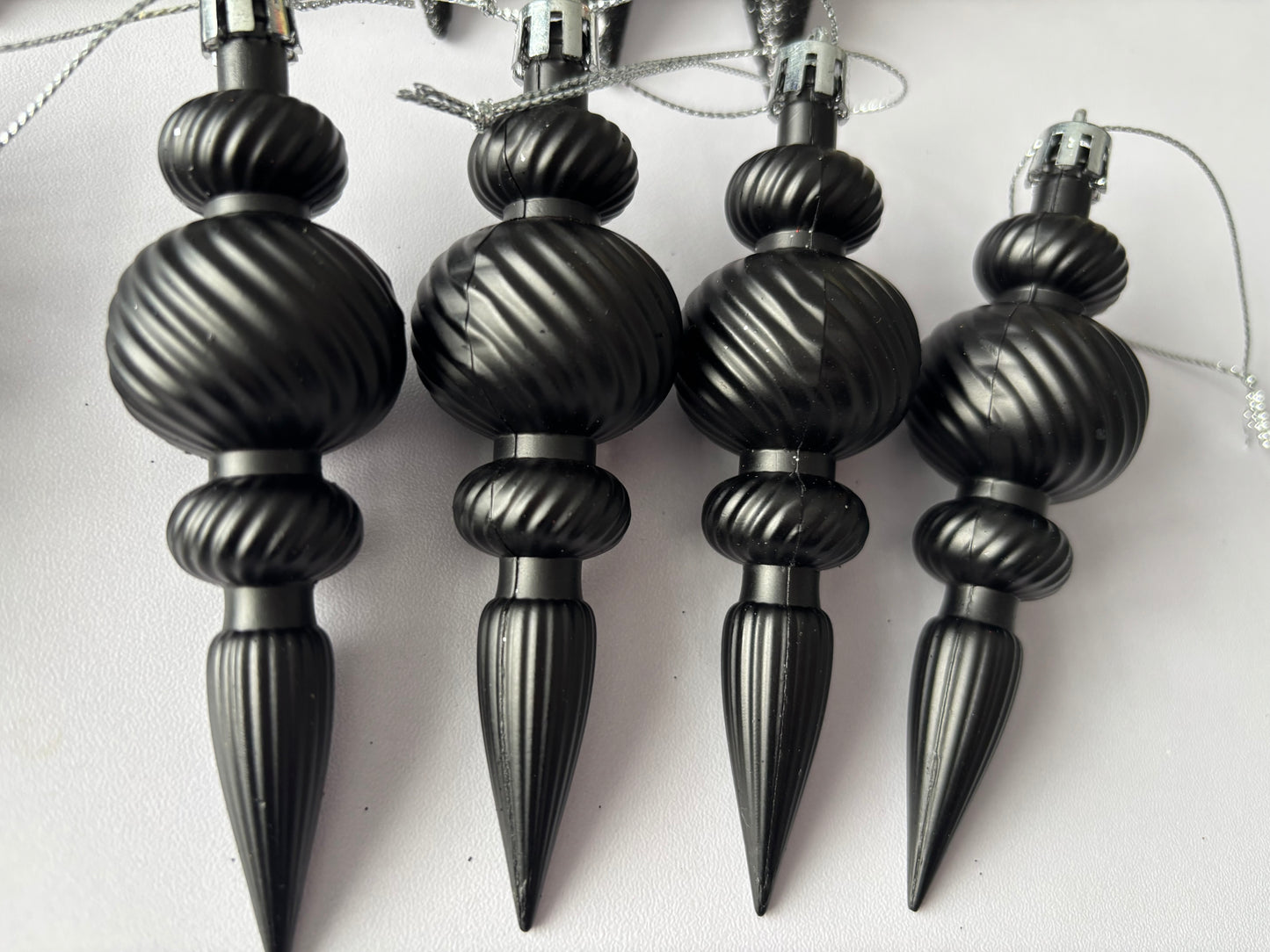 Close up of four black matte Christmas icicle finial baubles with large ribbed spherical tops, twisted ribbed middles and elongated pointed drop bases hanging from silver strings on a plain white background