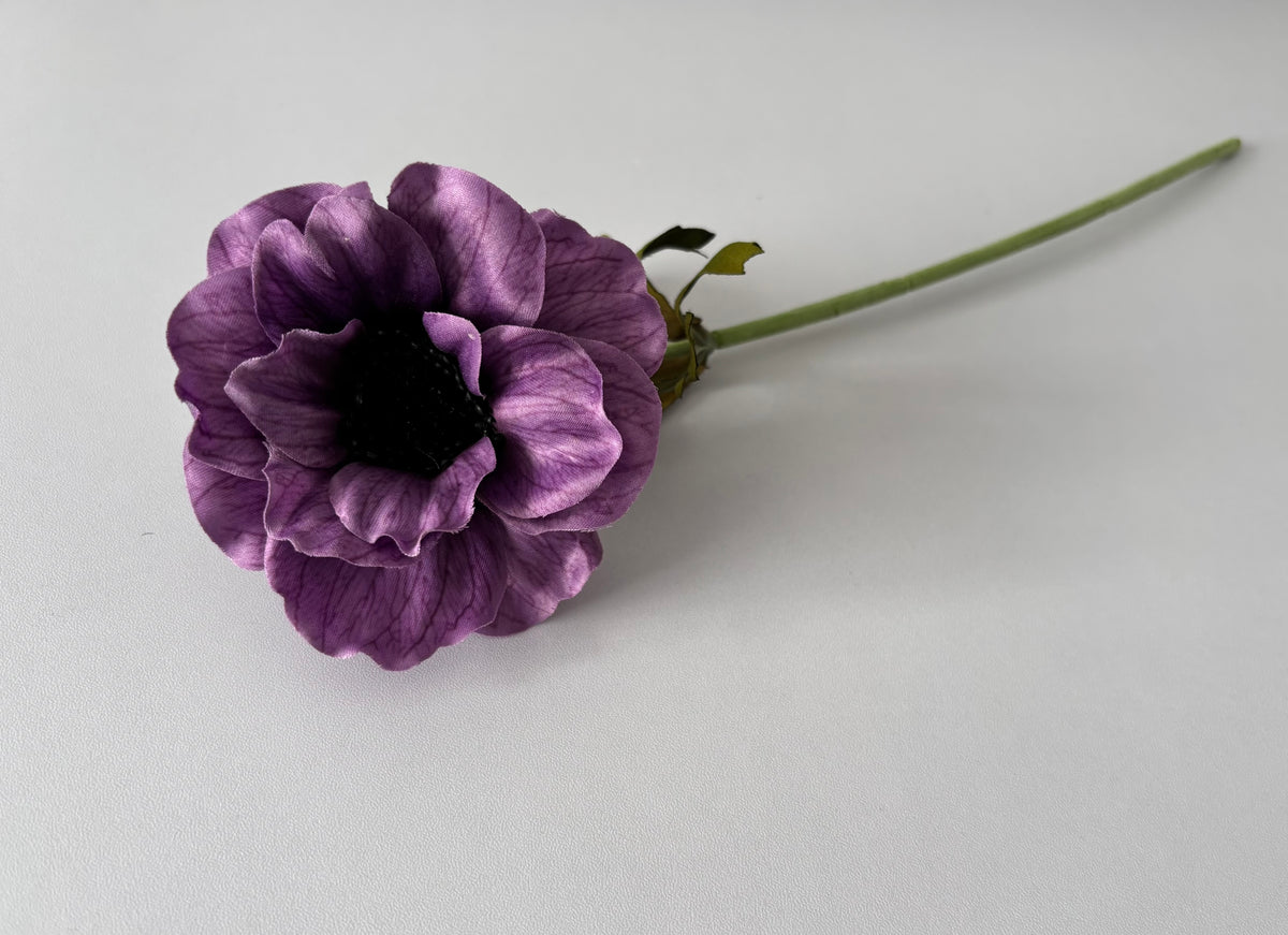 Upright portrait shot of an artificial purple silk anemone single stem held vertically showing the open layered cup-shaped flower head with ruffled purple petals, fine vein detail and a dramatic dark black raised centre with the fabric wrapped green stem and small green foliage leaves visible beneath against a plain light background