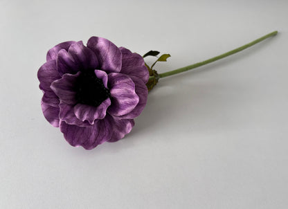 Upright portrait shot of an artificial purple silk anemone single stem held vertically showing the open layered cup-shaped flower head with ruffled purple petals, fine vein detail and a dramatic dark black raised centre with the fabric wrapped green stem and small green foliage leaves visible beneath against a plain light background