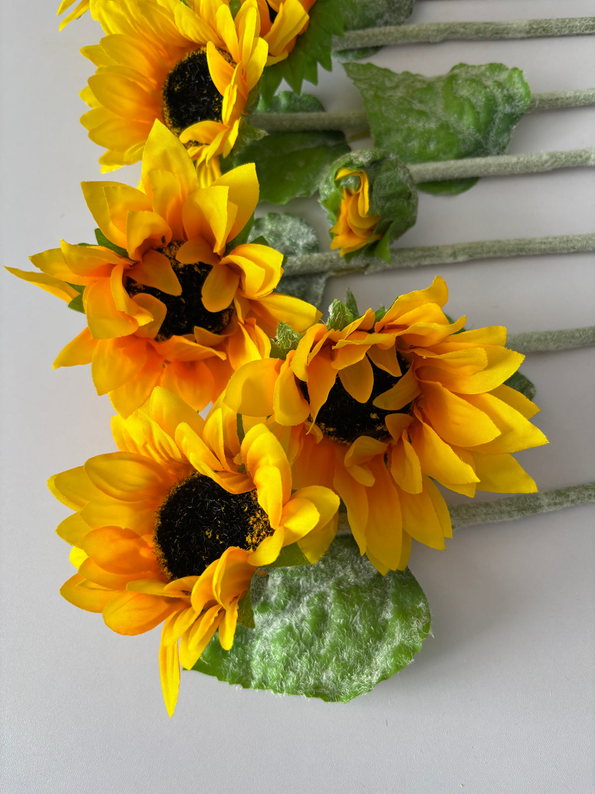 Close up portrait shot of three artificial sunflower stems showing the mixed size golden yellow flower heads with dark brown textured centres and lush bright green foliage with glittery silver textured stems on a plain light grey background