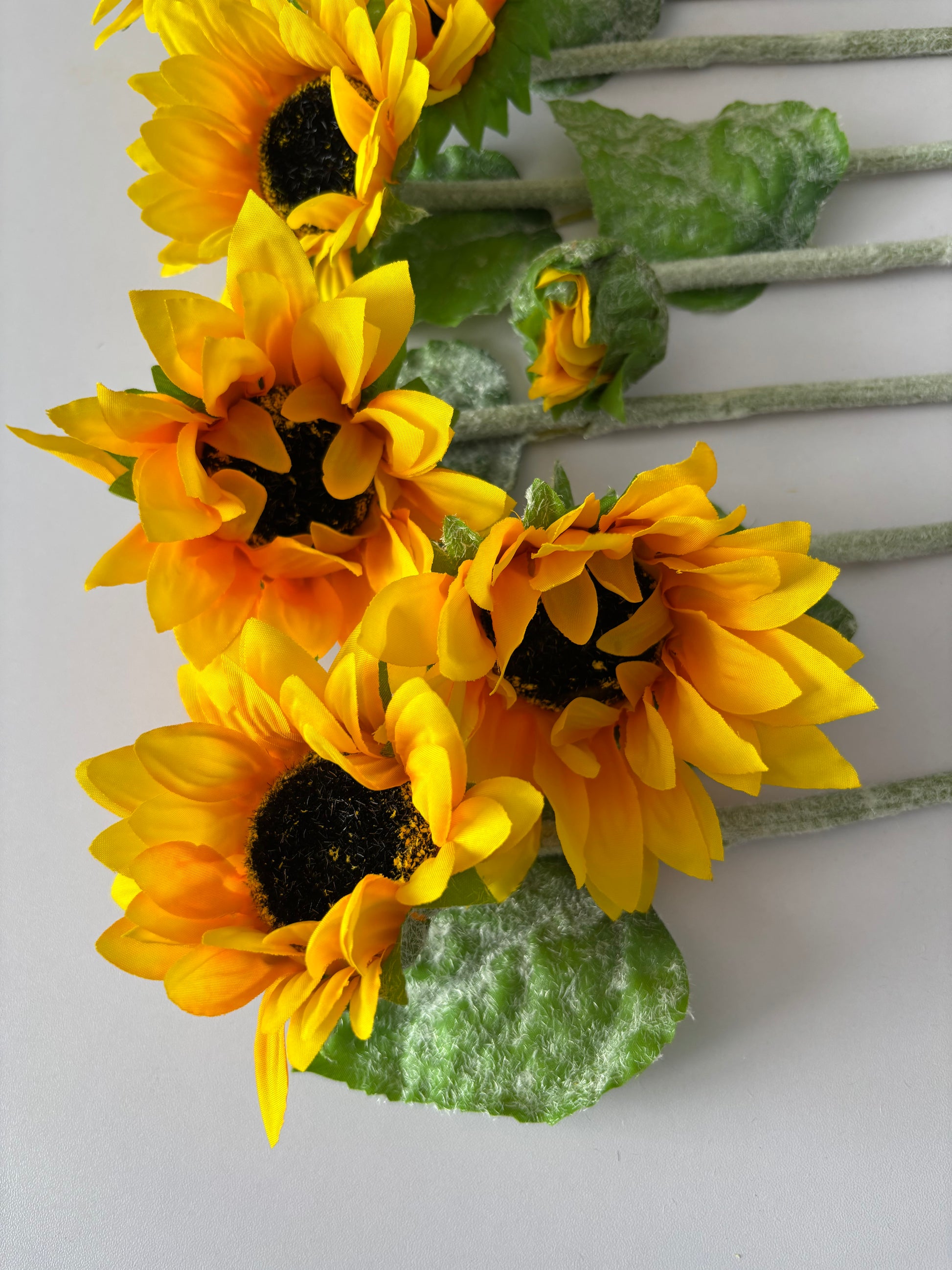 Close up portrait shot of three artificial sunflower stems showing the mixed size golden yellow flower heads with dark brown textured centres and lush bright green foliage with glittery silver textured stems on a plain light grey background
