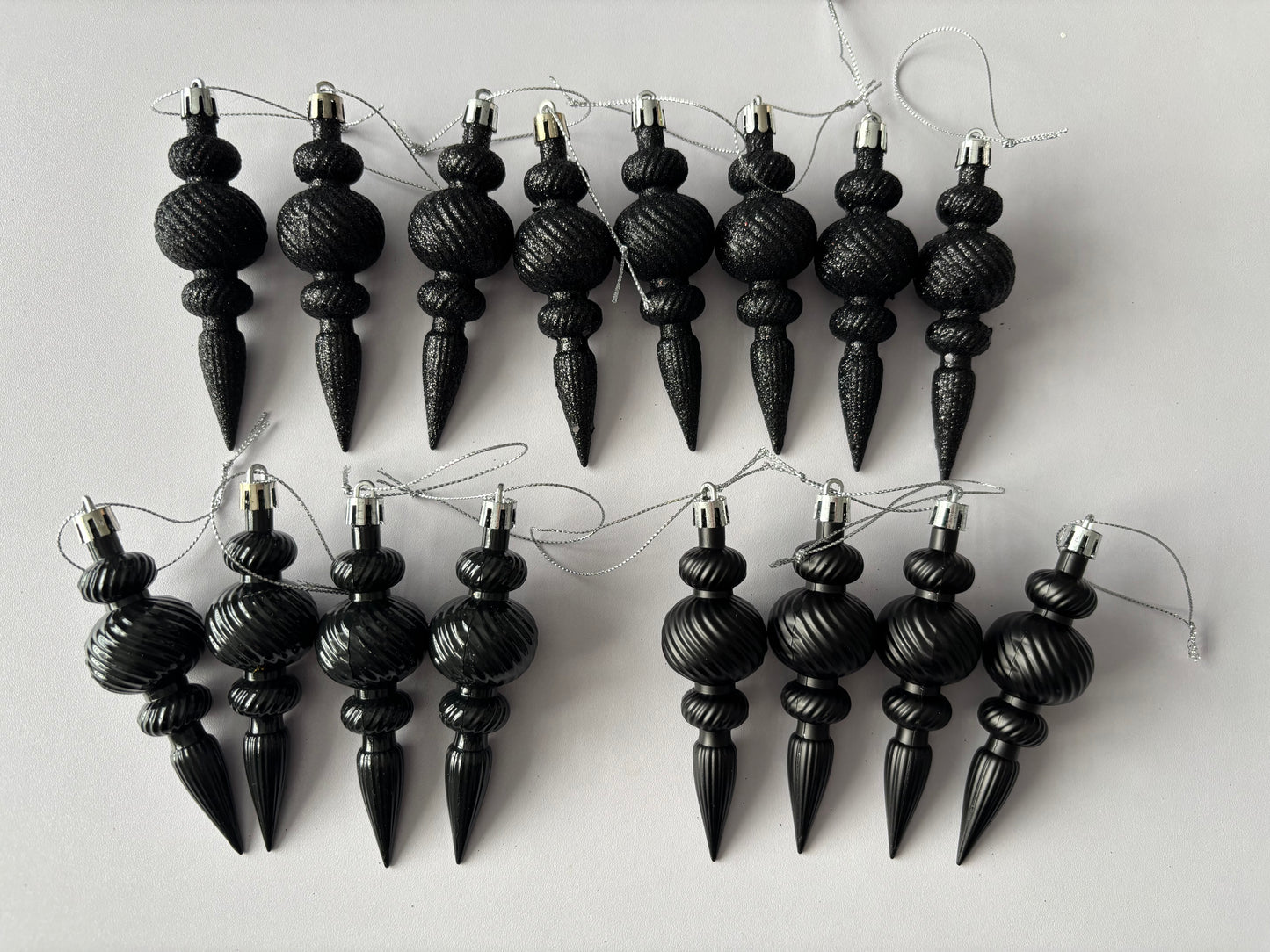 Flat lay of twelve black Christmas icicle finial baubles in glittered and matte finishes with ribbed spherical tops, beaded middles and elongated pointed drop bases arranged in two rows with silver hanging strings on a light grey background