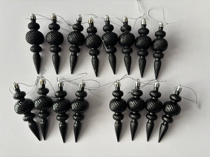 Flat lay of twelve black Christmas icicle finial baubles in glittered and matte finishes with ribbed spherical tops, beaded middles and elongated pointed drop bases arranged in two rows with silver hanging strings on a light grey background