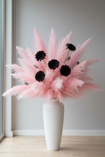 Artificial pink pampas sprays mixed with tall artificial black daisy stems arranged in a tall white ceramic floor vase on a light wood floor beside a window against a neutral grey wall