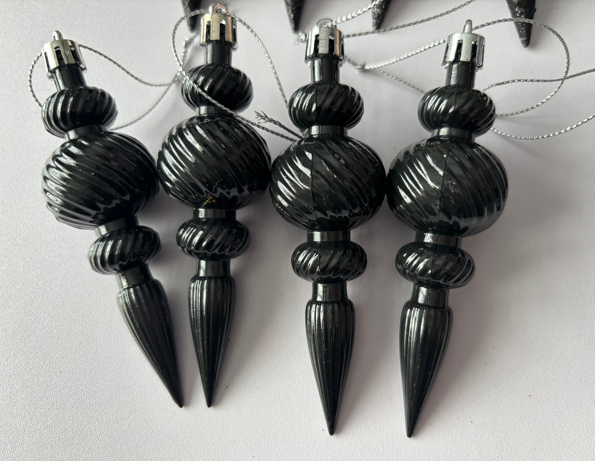 Close up of four black metallic Christmas icicle finial baubles with ribbed spherical tops, twisted ribbed middles and elongated pointed drop bases hanging from silver strings on a plain white background