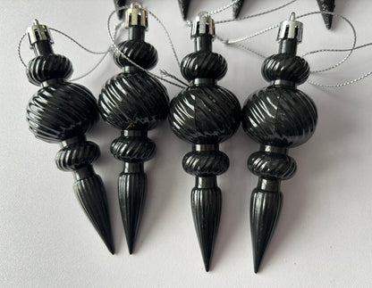 Close up of four black metallic Christmas icicle finial baubles with ribbed spherical tops, twisted ribbed middles and elongated pointed drop bases hanging from silver strings on a plain white background
