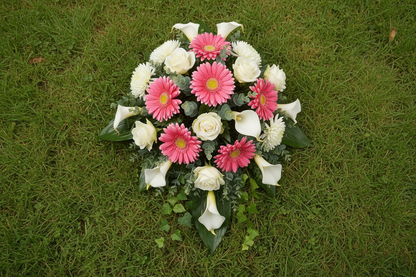 Artificial pink gerbera daisies styled in a circular funeral tribute with white roses, white calla lilies, green foliage and eucalyptus leaves placed on a green grass lawn