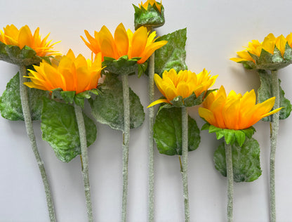 Seven artificial sunflower stems stood upright showing the mixed size golden yellow and orange flower heads with dark brown centres lush green foliage and glittery silver fuzzy textured stems on a plain light grey background