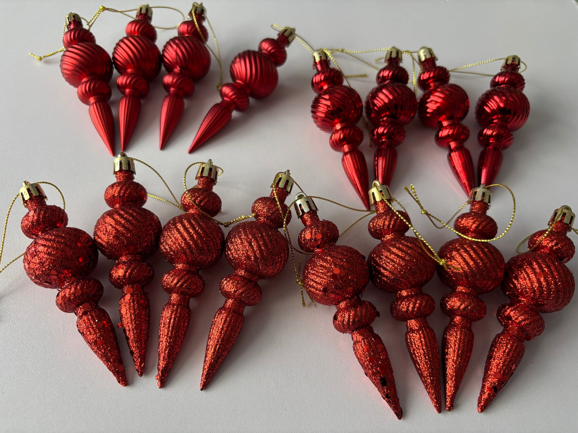 Flat lay of all 16 red Christmas spindle baubles in metallic and glittered finishes showing two distinct rows with ribbed spherical tops, beaded middles and elongated pointed drop bases with gold hanging strings on a plain white background