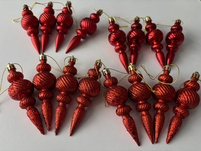 Flat lay of all 16 red Christmas spindle baubles in metallic and glittered finishes showing two distinct rows with ribbed spherical tops, beaded middles and elongated pointed drop bases with gold hanging strings on a plain white background