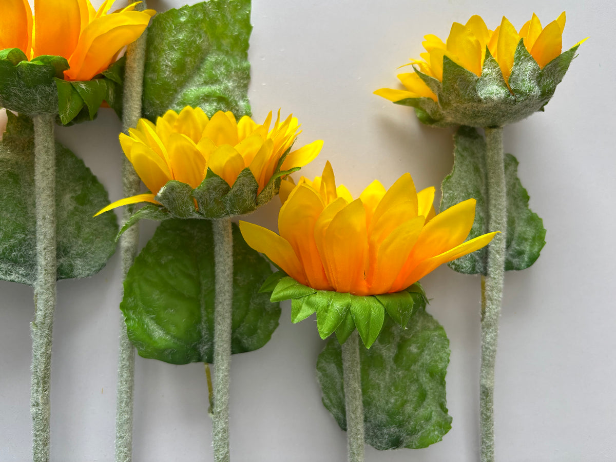 Four artificial sunflower stems stood upright showing the mixed size golden yellow and orange flower heads at various stages of bloom including one closed bud with lush green foliage and glittery silver textured stems on a plain light grey background