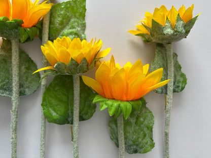 Four artificial sunflower stems stood upright showing the mixed size golden yellow and orange flower heads at various stages of bloom including one closed bud with lush green foliage and glittery silver textured stems on a plain light grey background