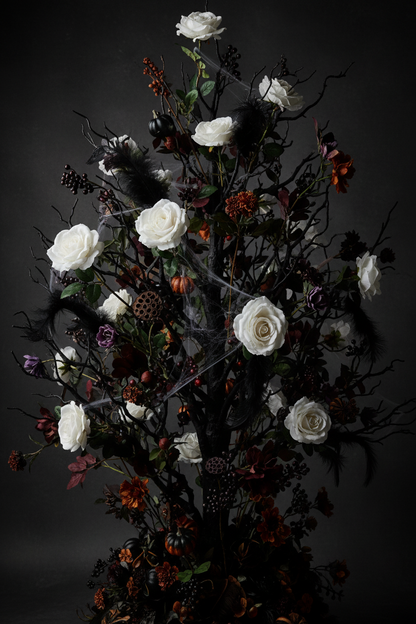 Artificial white velvet touch roses styled in a dramatic gothic Halloween floral arrangement with dark twisted bare branches, deep burgundy foliage, dark berries, purple blooms and orange berry accents against a dark black studio background