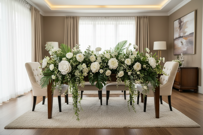 Artificial white velvet touch roses styled in a lush cascading wedding table centrepiece runner with white hydrangeas, tropical palm leaves, trailing green ivy and eucalyptus foliage along a formal dark wood dining table with cream upholstered chairs in an elegant neutral beige dining room