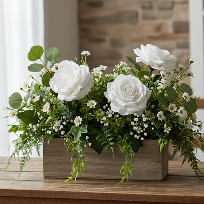 Artificial white velvet touch roses styled in a rustic wooden box centrepiece with green eucalyptus, trailing ferns, white gypsophila and small white daisy flowers on a natural wooden surface against a stone wall background