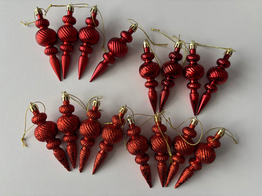 Flat lay of all 16 red Christmas spindle baubles in matte, metallic and glittered finishes with ribbed spherical tops, beaded middles and elongated pointed drop bases with gold hanging strings arranged in two rows on a plain white background