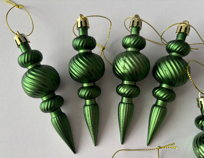 Close up of five green metallic Christmas spindle baubles with ribbed spherical tops, twisted ribbed middles and elongated pointed drop bases hanging from gold strings arranged on a plain light grey background
